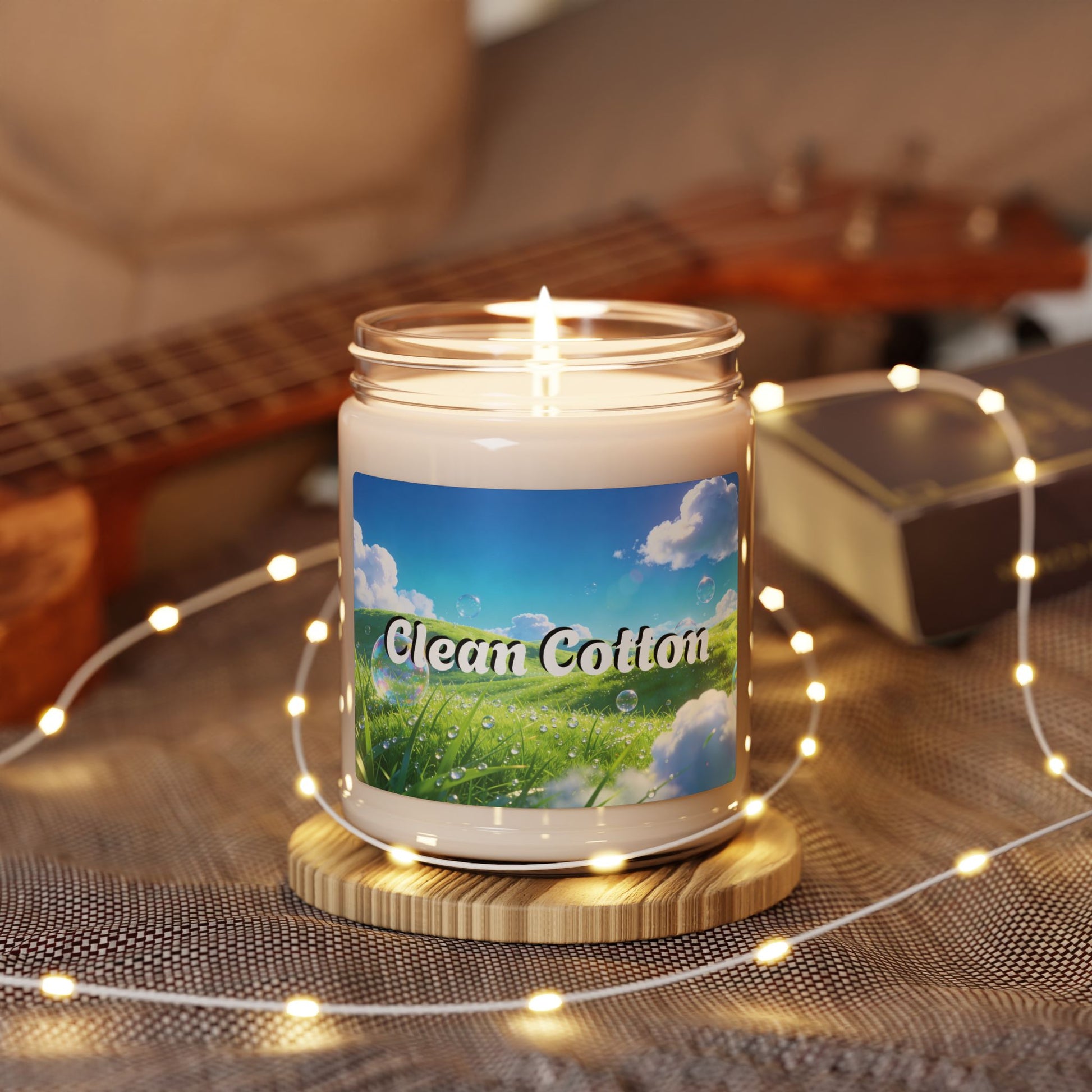 Candle with a 'Clean Cotton' label on a wooden coaster surrounded by string lights, with a guitar and box in the background.